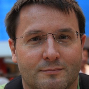 Head-and-shoulders photograph of Kevin Thomas Luzbetak: short dark hair, thin-rimmed glasses, looking straight at the camera against a blurred outdoor background.